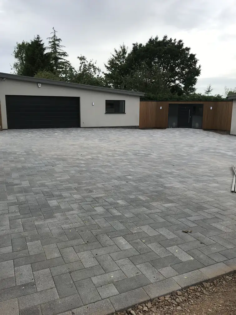 Expansive block paved forecourt and driveway fronting a rendered double garage with cedar-clad entrance.