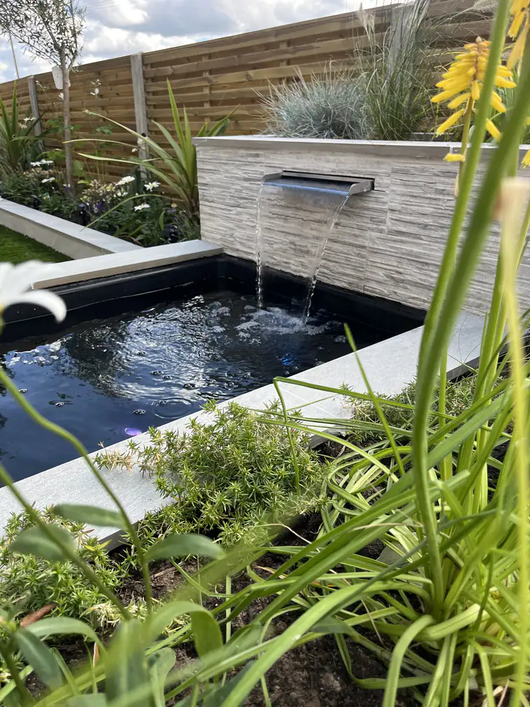 Cascading water blade into a dark pond framed by fescues and alpine planting, movement and calm in one scene.