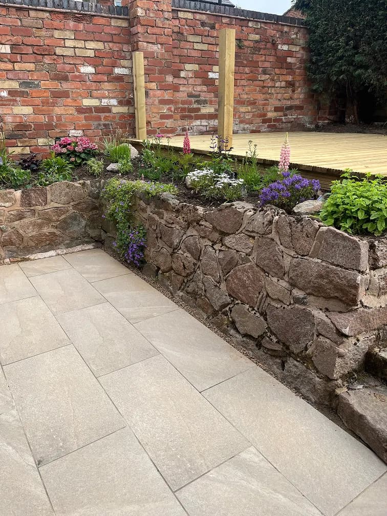 Raised stone flower bed with colourful planting, porcelain patio paving, brick wall and timber deck area.