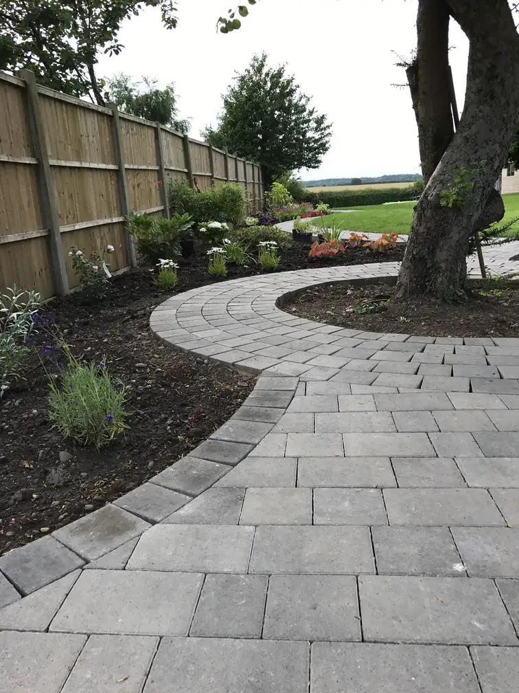 Block-paved garden path curving around a mature tree with mulched planting beds beside a new closeboard fence.