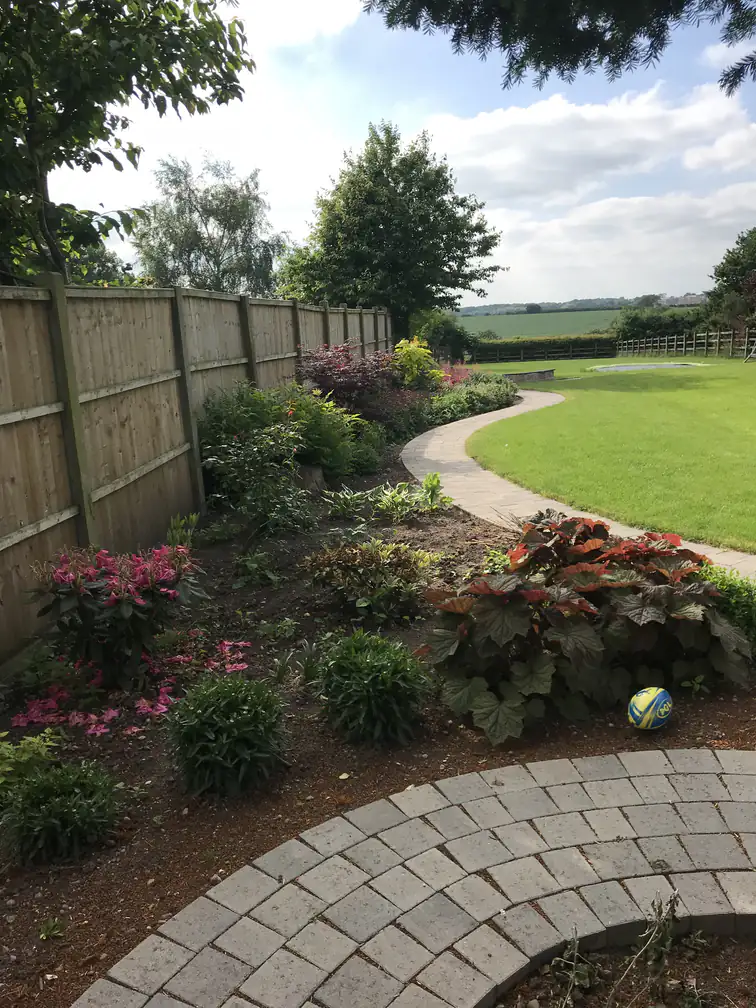 Curved block path threading between colourful flower borders and lawn beside new closeboard fencing.