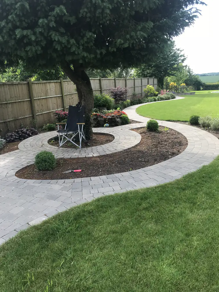 Spiral block-paved path circling a specimen tree with fresh planting, a playful focal point in a family lawn.