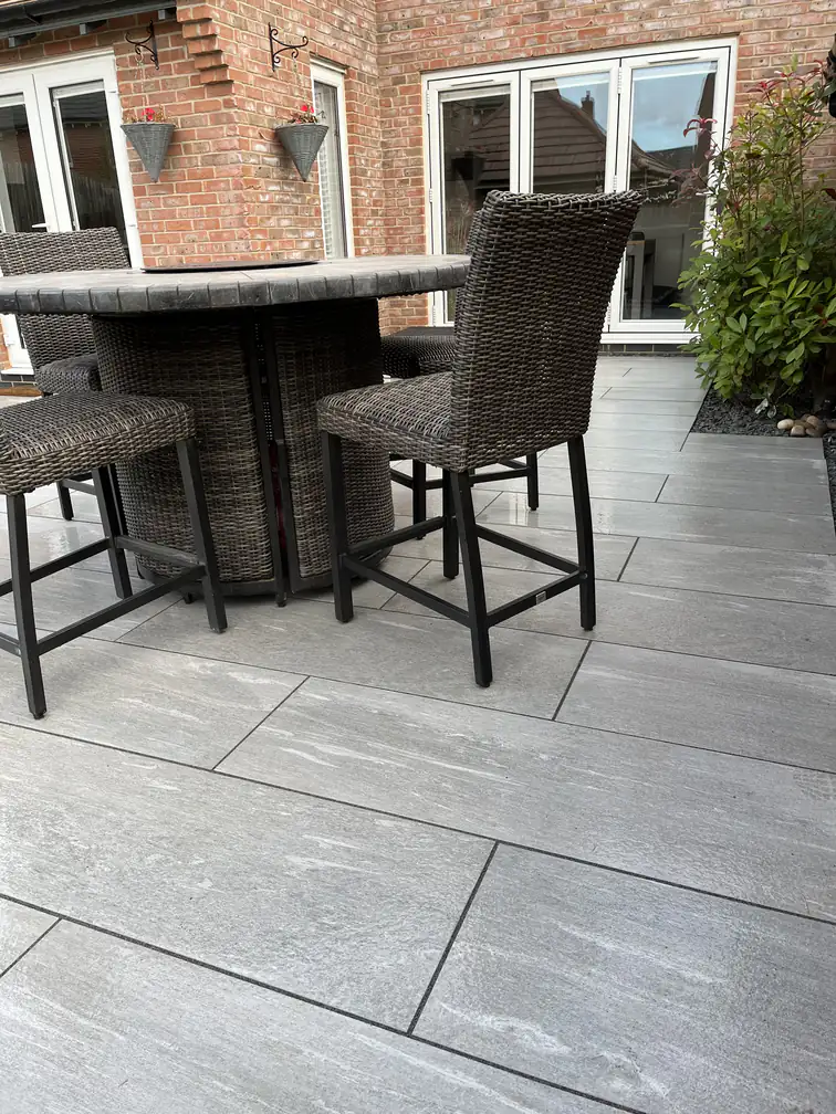 Rattan bar table and stools on a silver porcelain terrace against warm brickwork, bistro dining at home.