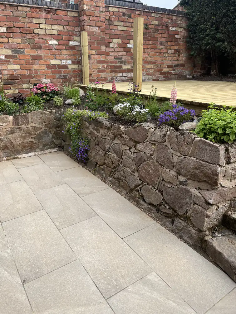 Garden landscaping with a reclaimed stone raised bed full of cottage planting beside new timber decking in a walled garden.