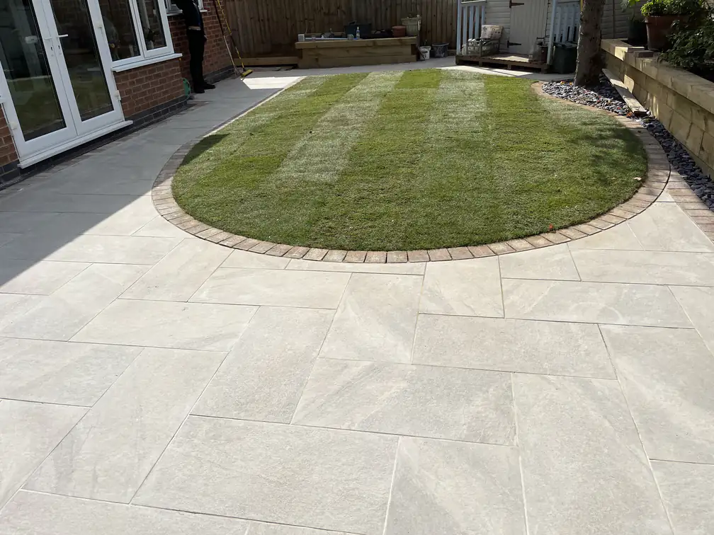 Oval lawn ringed with brindle sett edging beside a white porcelain patio and sleeper retaining wall.