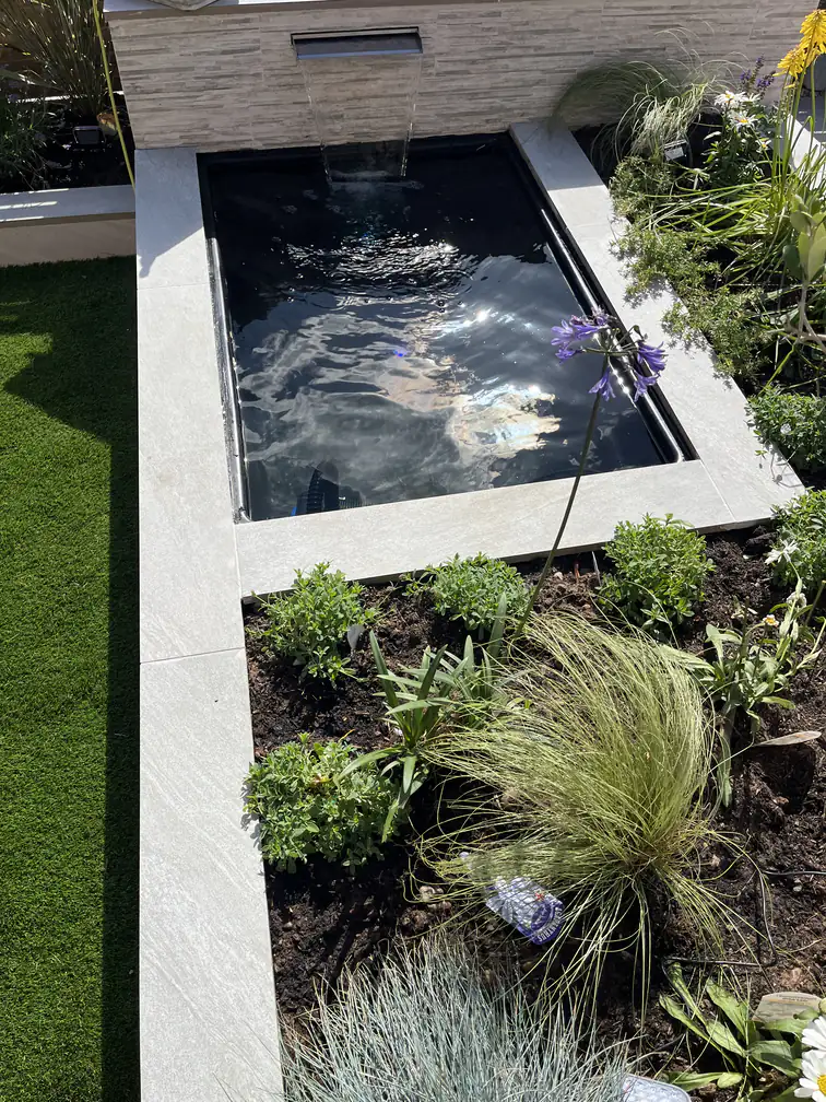 Porcelain raised pond with stainless water blade surrounded by artificial lawn and fresh planting in a contemporary garden.