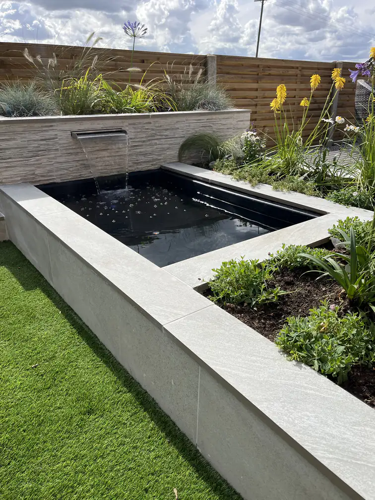 Raised pond with waterfall blade beside pristine artificial lawn, the centrepiece of a low-maintenance modern garden.