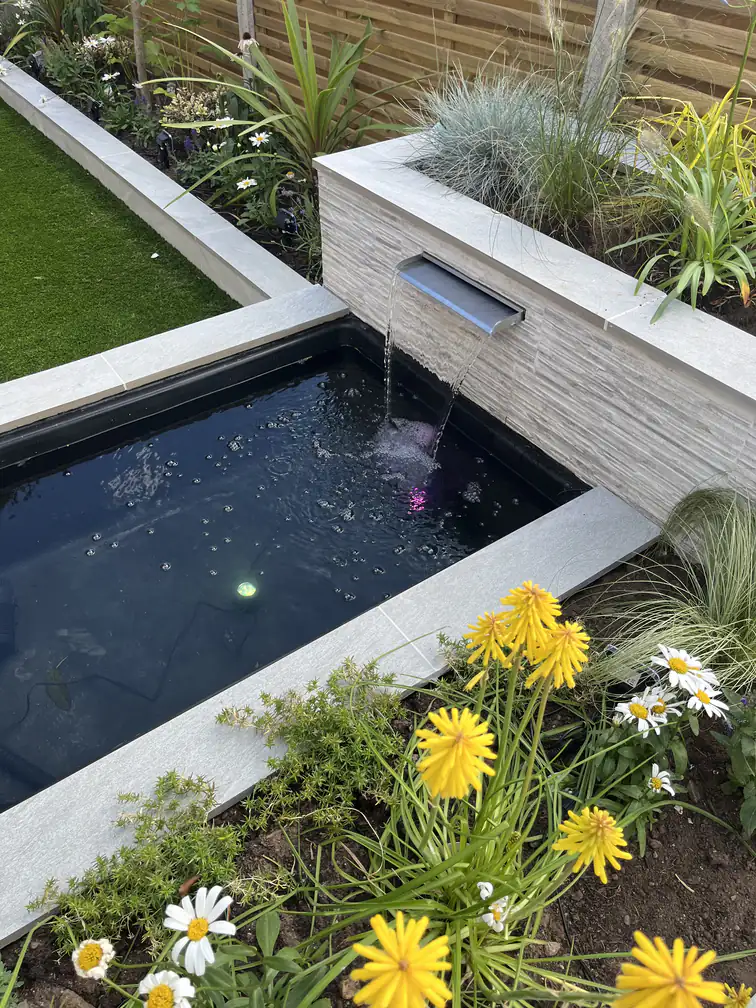 Reflective black pond edged with corn marigolds and daisies, vivid low-maintenance colour against porcelain stone.