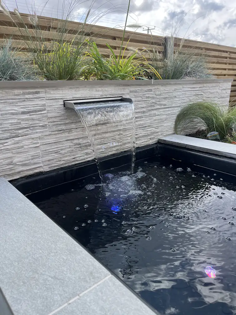Stone-clad waterfall blade cascading into a dark reflection pond with underwater lighting, a dramatic evening feature.