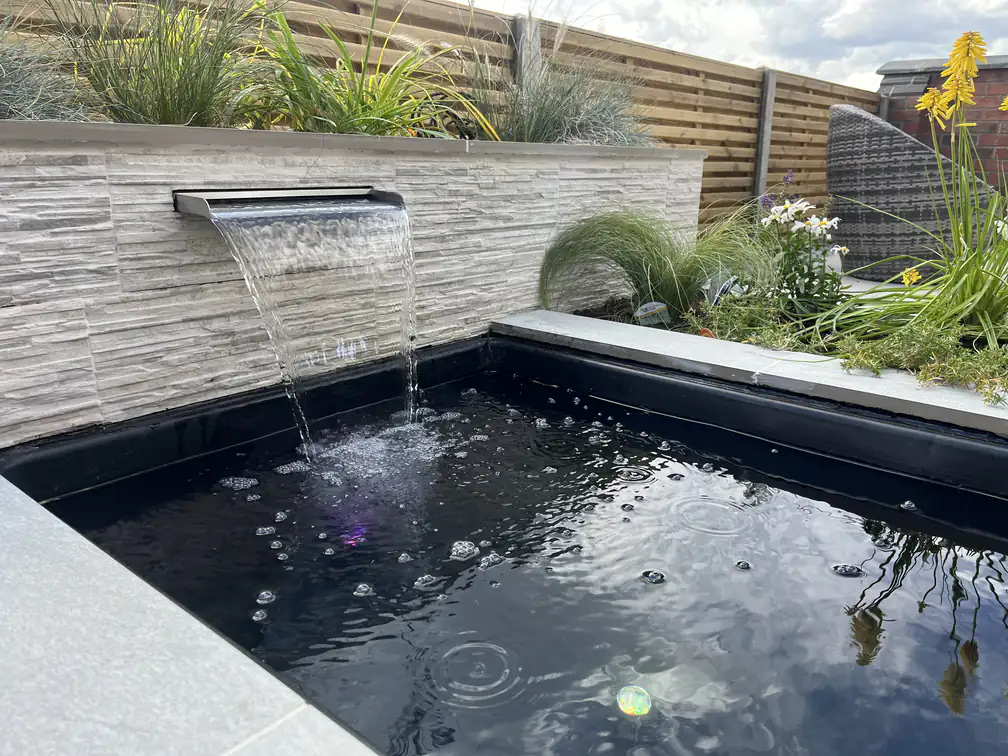 Textured stone water wall with twin cascades into a raised pond, framed by ornamental grasses and red hot pokers.