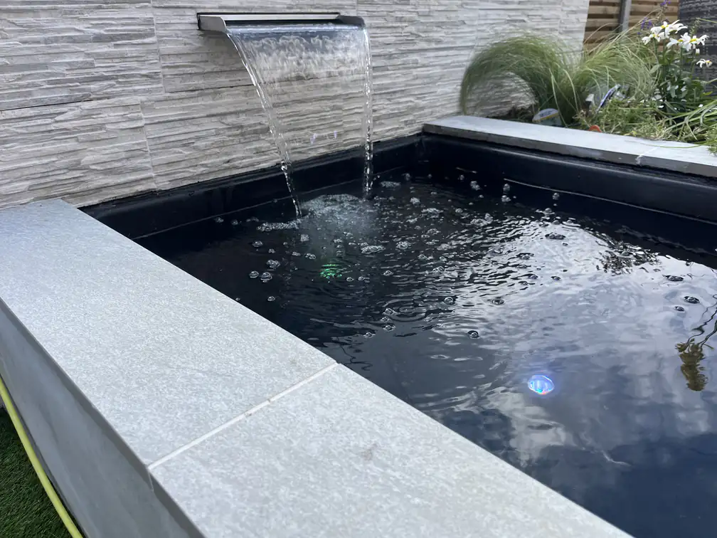 Close-up of a stainless waterfall blade feeding a black reflection pool, crisp water movement over natural stone cladding.